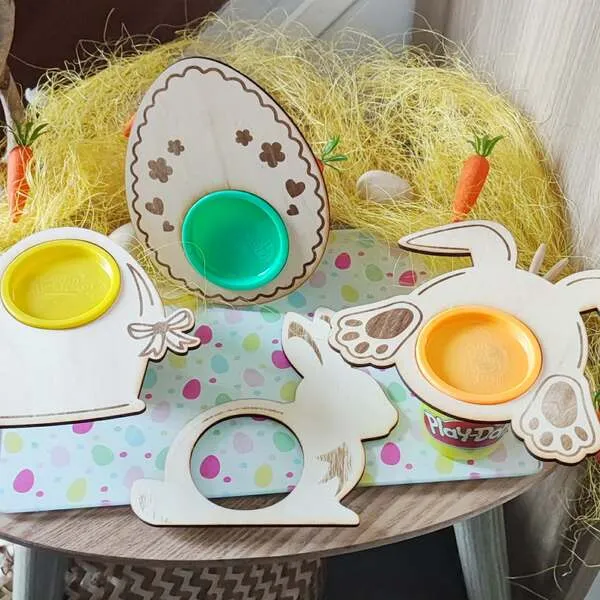 Supports en bois thème Pâques pour pots de pâte à modeler, formes œuf et lapin avec emplacements ronds colorés, posés sur une table décorée façon chasse aux œufs.