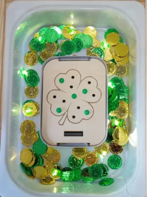 Insert trèfle en bois placé dans une table sensorielle avec pièces d’or vertes et dorées thème Saint Patrick.