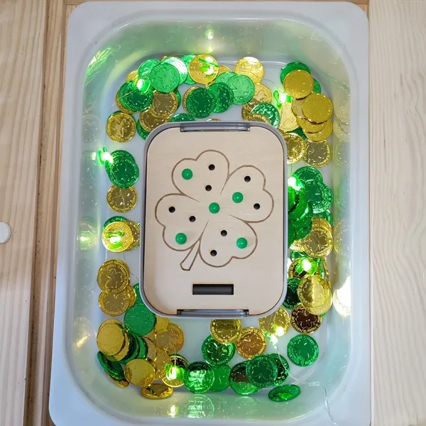 Insert trèfle en bois placé dans une table sensorielle avec pièces d’or vertes et dorées thème Saint Patrick.
