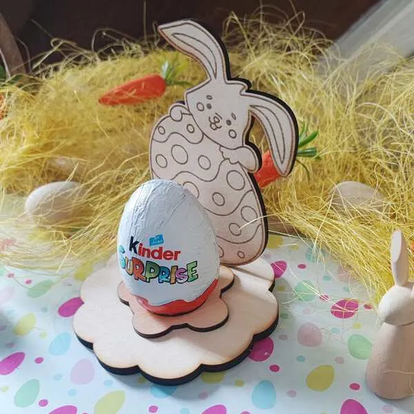 Support en bois pour œuf de Pâques avec lapin appuyé sur un grand œuf décoré, socle en forme de fleur et œuf Kinder Surprise au centre.