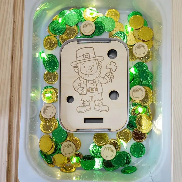 Insert en bois gravé lutin Saint Patrick dans bac sensoriel avec pièces dorées et vertes.