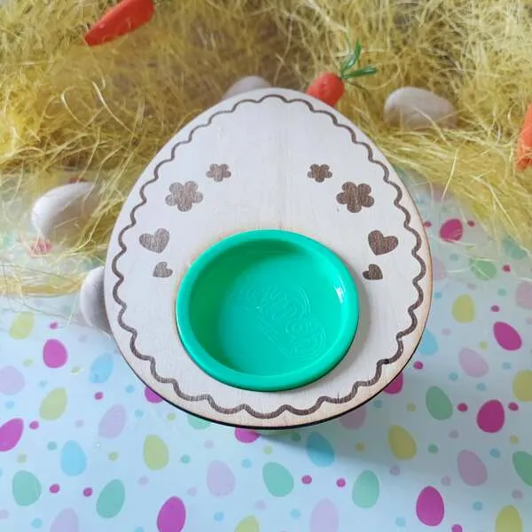 Support en bois en forme d’œuf de Pâques avec gravures décoratives (cœurs et fleurs) et emplacement central pour pot de pâte à modeler vert.