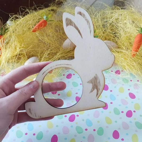Anneau en bois en forme de lapin avec ouverture ronde pour pot de pâte à modeler, tenu en main sur une table décorée pour Pâques.