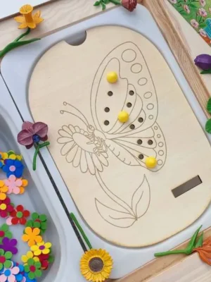 Insert papillon en bois pour table Flisat avec activité de placement de pions et bac rempli de fleurs colorées.