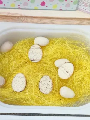 Bac sensoriel Pâques avec paille jaune et œufs en bois décorés.