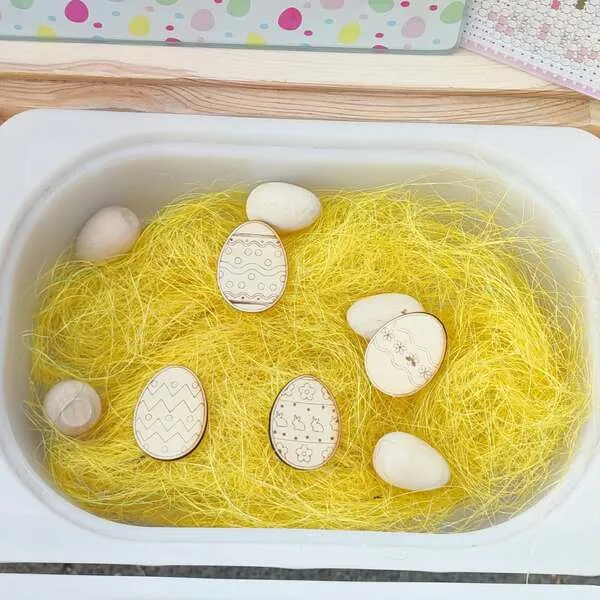 Bac sensoriel Pâques avec paille jaune et œufs en bois décorés.