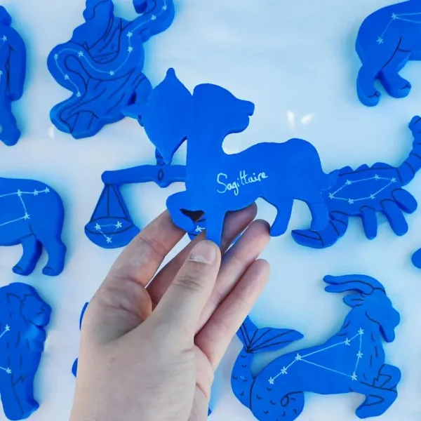 Main tenant une figurine en bois du signe Sagittaire, peinte en bleu, avec son nom inscrit au dos, devant une collection de signes astrologiques et constellations.
