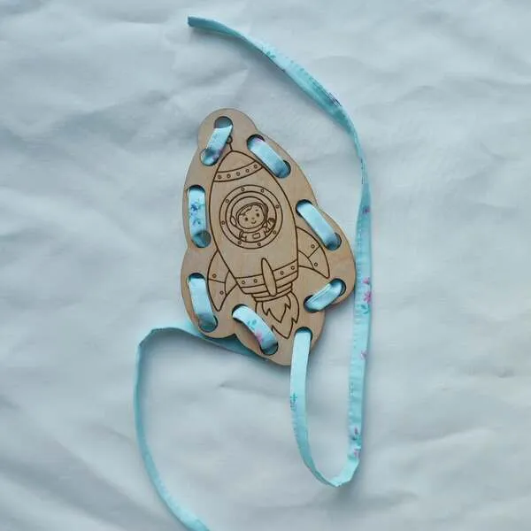 Carte à lacets en bois représentant une fusée avec un jeune astronaute dans le hublot et un ruban bleu à enfiler.