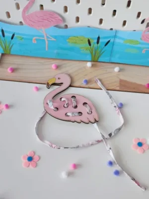 Carte à lacer Flamant rose en bois avec un lacet en tissu, présentée dans une mise en scène tropicale avec un décor de lagon, des flamants roses et de petits éléments décoratifs roses et violets.
