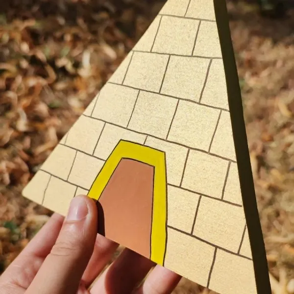 Pyramide en bois peinte à la main, issue du mini monde Égypte, présentée en gros plan avec une main la tenant en extérieur.