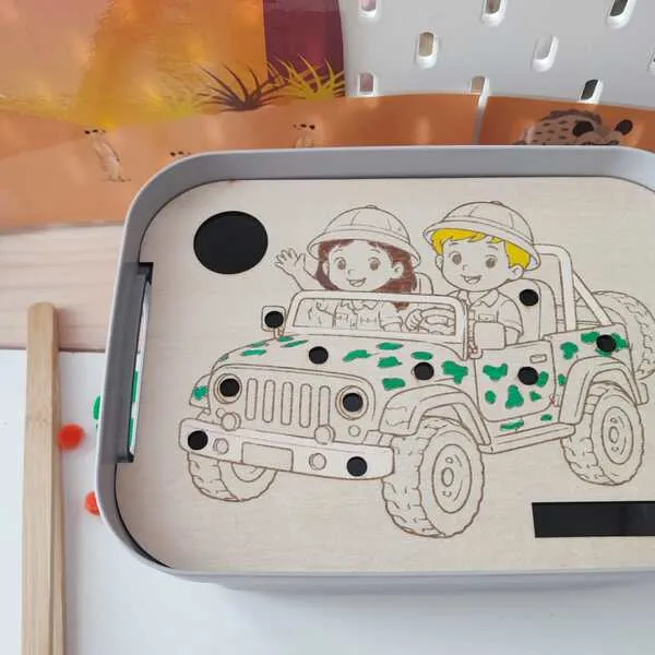 Insert en bois pour bac sensoriel LIVV 2L sur le thème de la savane. Cette activité de motricité fine représente deux enfants en voiture de safari. Les enfants utilisent une pince pour placer les pompons dans les différents emplacements du véhicule, favorisant la coordination œil-main, la précision des gestes et la concentration.