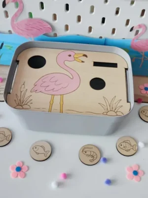 Insert Livv 2L en bois représentant un flamant rose avec le bec ouvert. Des jetons illustrés de poissons et de crevettes sont disposés autour du bac, prêts à être insérés pour nourrir l'oiseau.