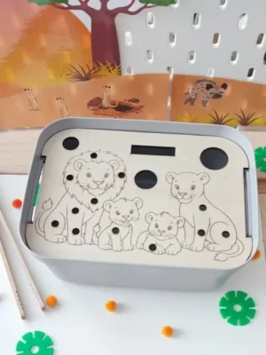 Insert éducatif en bois pour bac sensoriel LIVV 2L sur le thème de la savane représentant une famille de lions. L'enfant utilise une pince pour placer des pompons dans les emplacements du lion, de la lionne et des lionceaux afin de développer la motricité fine, la coordination œil-main, la précision des gestes et la concentration. Activité Montessori idéale pour les assistantes maternelles, MAM, crèches, écoles maternelles et familles.