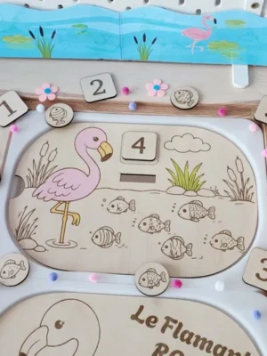 Insert Livv 2L en bois représentant un flamant rose entouré de poissons. L'enfant compte les poissons visibles dans l'étang, sélectionne le bon chiffre de 1 à 6 et l'insère dans l'encoche prévue.