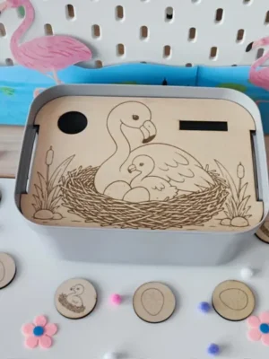 Insert Livv 2L en bois représentant un nid de flamants roses avec un adulte, un bébé et des œufs. Des jetons illustrés d'œufs et de bébés flamants sont disposés autour du bac pour compléter l'activité.