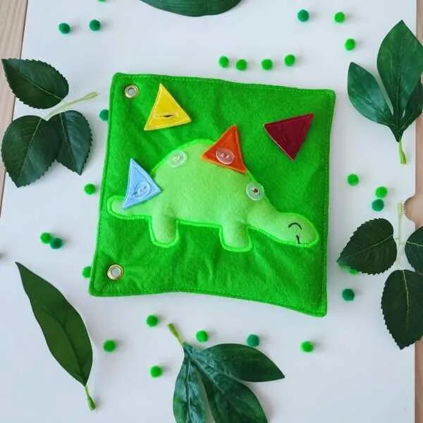 Page de Quiet Book en feutrine représentant un stégosaure avec des plaques dorsales triangulaires colorées à boutonner pour travailler les formes et la motricité fine.