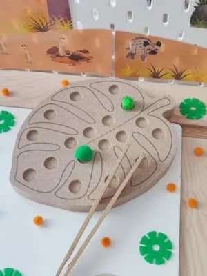 Plateau de motricité fine en bois en forme de feuille tropicale, sur le thème de la savane. L'enfant utilise une pince pour déplacer et placer des pompons dans les différents creux du plateau, développant sa précision, sa coordination œil-main et sa concentration.