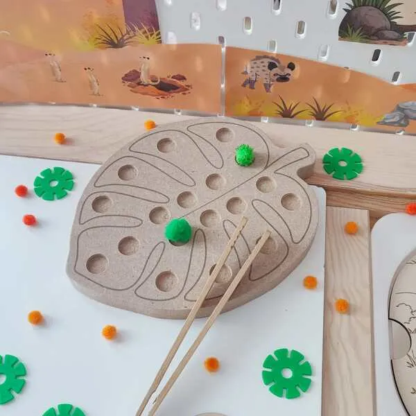 Plateau de motricité fine en bois en forme de feuille tropicale, sur le thème de la savane. L'enfant utilise une pince pour déplacer et placer des pompons dans les différents creux du plateau, développant sa précision, sa coordination œil-main et sa concentration.