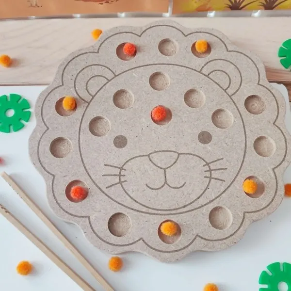 Gros plan d'un plateau de motricité fine en MDF en forme de tête de lion, avec plusieurs cavités contenant des pompons orange et jaunes. Une pince en bois permet de manipuler les pompons pour développer la motricité fine, la coordination œil-main et la concentration des jeunes enfants.