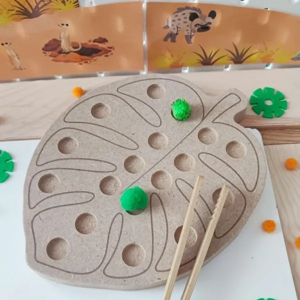Plateau de motricité fine en bois en forme de feuille de monstera avec plusieurs emplacements pour déplacer des pompons à l'aide d'une pince. Cette activité, présentée dans un univers sur le thème de la savane, favorise la précision des gestes, la coordination œil-main et la concentration des jeunes enfants.