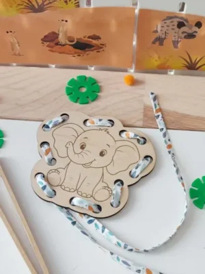 Carte à lacer en bois représentant un éléphant souriant avec un lacet épais passé dans les trous. Cette activité développe la motricité fine, la coordination œil-main, la précision des gestes et prépare aux premiers apprentissages de l'écriture.
