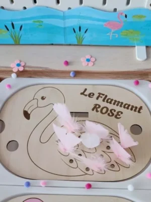 Insert Trofast en bois représentant un flamant rose avec plusieurs plumes roses déjà insérées, présenté dans un décor inspiré d'un lagon.