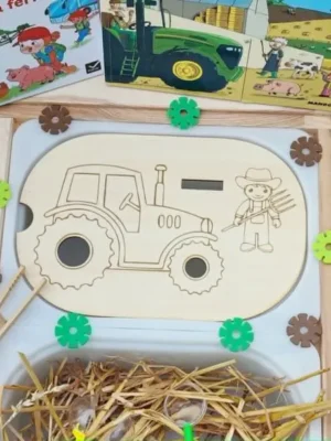 Insert en bois pour bac Trofast représentant un tracteur et un fermier, accompagné de jetons à placer avec une pince. Le support est présenté dans un bac sensoriel avec de la paille, des engrenages et des livres sur le thème de la ferme.