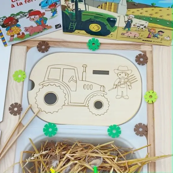 Insert en bois pour bac Trofast représentant un tracteur et un fermier, accompagné de jetons à placer avec une pince. Le support est présenté dans un bac sensoriel avec de la paille, des engrenages et des livres sur le thème de la ferme.