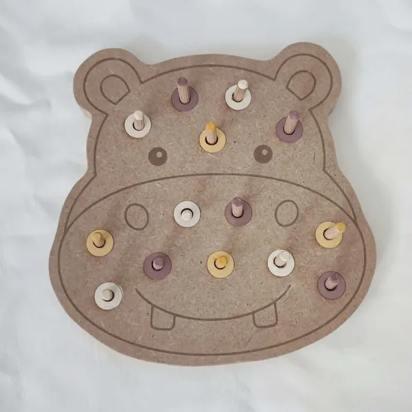 Plateau à chevilles en MDF en forme d'hippopotame avec des chevilles en bois et des anneaux de trois couleurs. Cette activité éducative permet de développer la motricité fine, le tri des couleurs, la coordination œil-main et la précision des gestes.