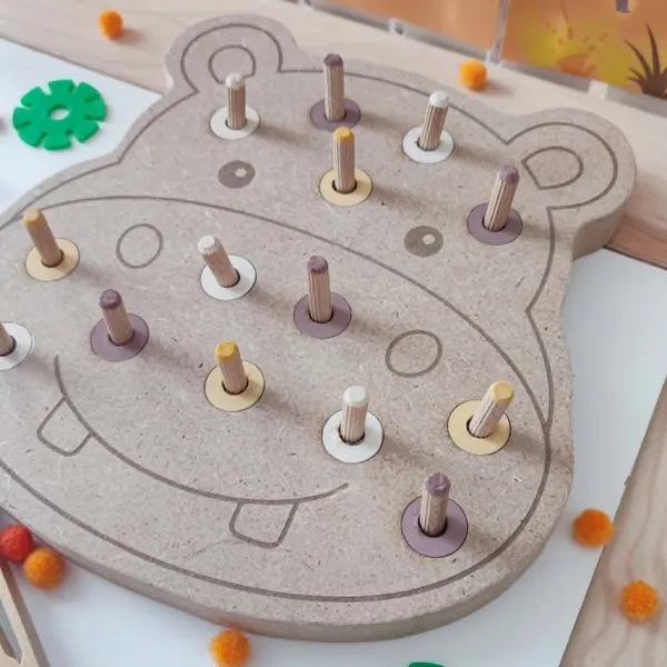 Gros plan d'un plateau à chevilles en MDF en forme d'hippopotame avec des chevilles en bois colorées. Cette activité de manipulation permet aux enfants de travailler la motricité fine, la coordination œil-main, le tri des couleurs et la précision des gestes.