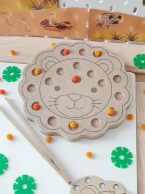 Plateau de motricité fine en MDF en forme de tête de lion avec plusieurs cavités remplies de pompons colorés. L'enfant utilise une pince pour déplacer les pompons, développant sa motricité fine, sa coordination œil-main et sa concentration dans un univers inspiré de la savane.