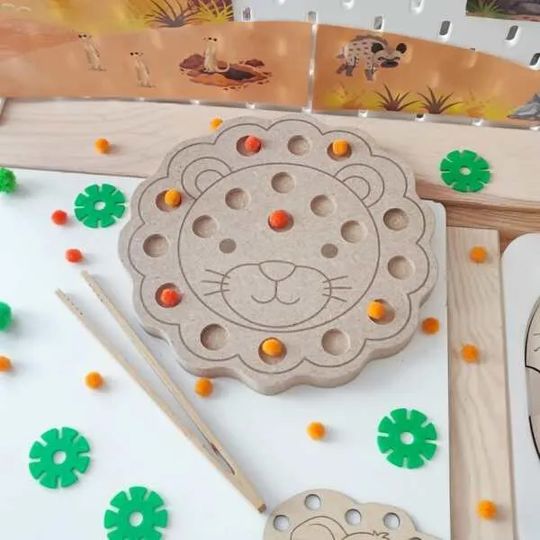 Plateau de motricité fine en MDF en forme de tête de lion avec plusieurs cavités remplies de pompons colorés. L'enfant utilise une pince pour déplacer les pompons, développant sa motricité fine, sa coordination œil-main et sa concentration dans un univers inspiré de la savane.