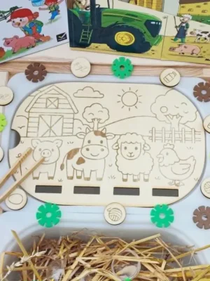 Insert en bois pour bac Trofast représentant une scène de ferme avec un cochon, une vache, un mouton et une poule. Des jetons illustrés sont à associer aux animaux. Le support est présenté dans un bac sensoriel avec de la paille.