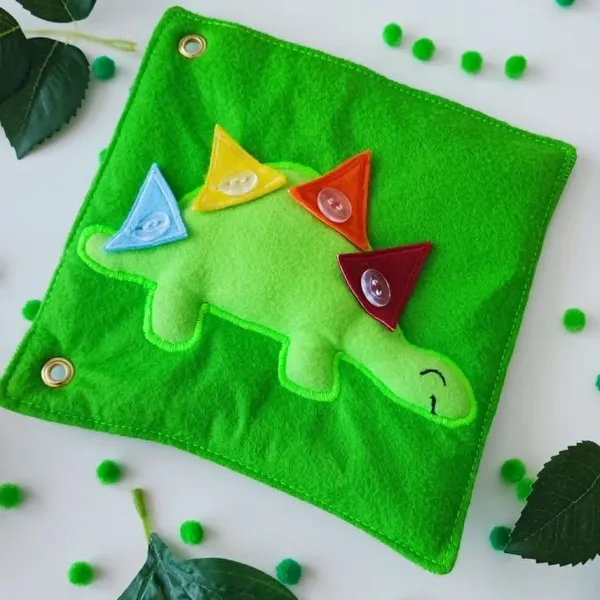Page de Quiet Book en feutrine représentant un stégosaure vert avec quatre plaques dorsales colorées à boutonner pour développer la motricité fine.