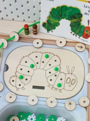 Plateau pédagogique en bois inspiré de La chenille qui fait des trous, avec une chenille gravée, des jetons illustrés d'aliments, des perles vertes à compter et une carte du livre en arrière-plan.