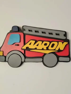 Plaque de porte personnalisée en bois représentant un camion de pompier rouge en relief, avec une échelle grise, des roues grises et le prénom Aaron en lettres jaunes.
