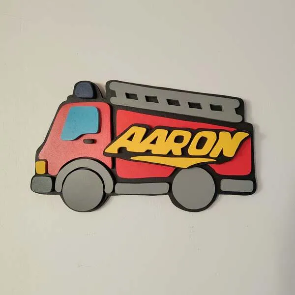 Plaque de porte personnalisée en bois représentant un camion de pompier rouge en relief, avec une échelle grise, des roues grises et le prénom Aaron en lettres jaunes.