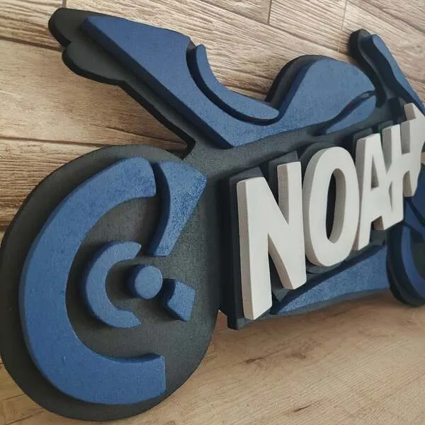 Gros plan d’une plaque de porte moto personnalisée Noah en bois, avec roue bleue et prénom blanc en relief sur fond noir.