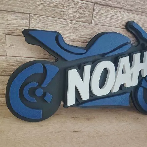 Gros plan sur une plaque de porte moto personnalisée en bois bleu et noir, avec le prénom Noah en lettres blanches en relief.