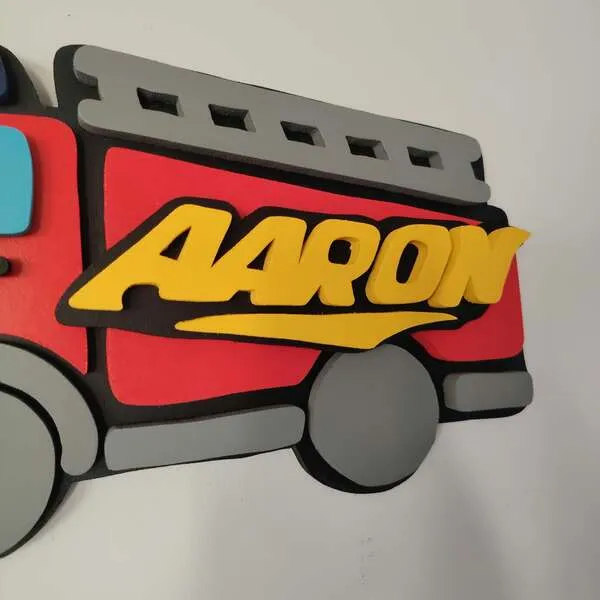 Gros plan sur le prénom Aaron en lettres jaunes en relief, fixé sur une plaque de porte camion de pompier rouge en bois avec échelle et roue grises.