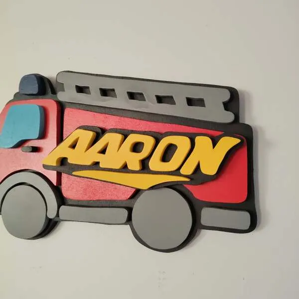 Gros plan sur une plaque de porte personnalisée en bois en forme de camion de pompier rouge, avec le prénom Aaron en lettres jaunes en relief, une échelle grise et des roues superposées.